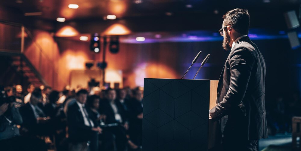 A man speaking in front of a crowd in a dim hall.