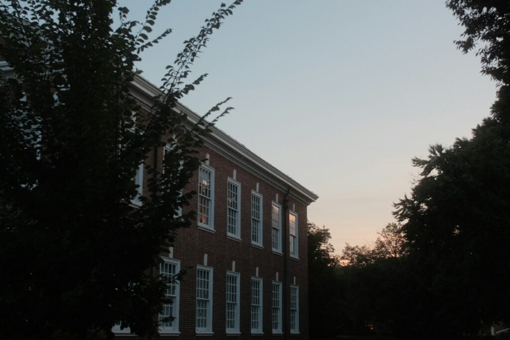 A low-angle view of a multi-story red brick colonial-style university building at dusk, framed by dark silhouettes of trees on both sides.