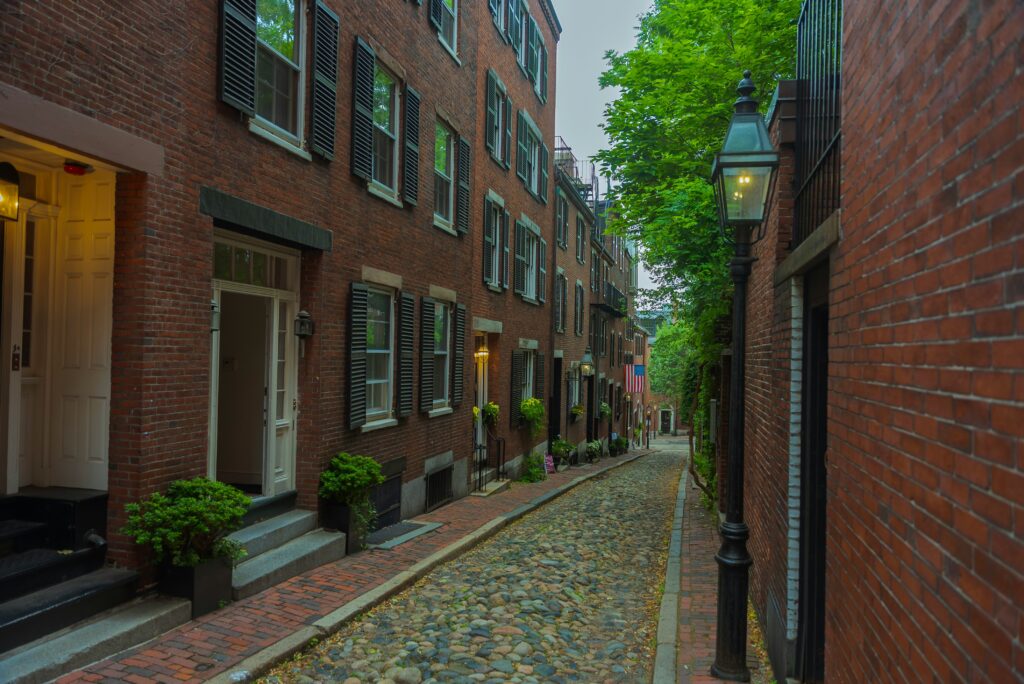 A cobblestone sidewalk in Massachusetts with a 3 story residential building on both sides a gas street lamp on the middle right side of the photo.
